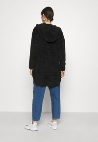 Kvinde med mørkt hår i en bundt iført sølv hoop øreringe, en sort hættefleecejakke, blå jeans og hvide sneakers, vender væk fra kameraet.