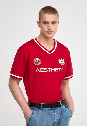 Jersey sportivo rosso con accenti bianchi, design a V, logo sul petto e testo audace "ESTETICO". Indossato con pantaloni corti in denim blu.