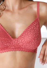 Bralette in pizzo rosa con forma triangolare, bordi scallop e spalline sottili. Un motivo floreale testurizzato ricopre la superficie.