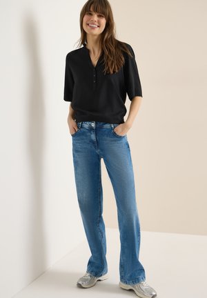 Femme souriante, les mains dans les poches, portant un chemisier noir à manches courtes, un jean bleu et des baskets argentées, debout devant un mur bicolore.