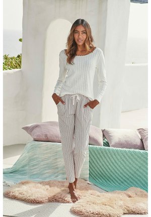 Maglietta bianca a maniche lunghe con texture abbinata a pantaloni leggeri a righe. Il modello è in piedi su un tappeto soffice con cuscini strutturati sullo sfondo.