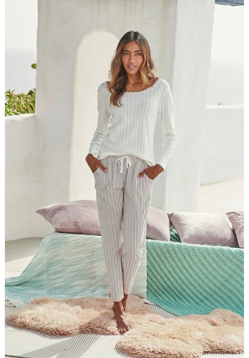 Maglietta bianca a maniche lunghe con texture abbinata a pantaloni leggeri a righe. Il modello è in piedi su un tappeto soffice con cuscini strutturati sullo sfondo.