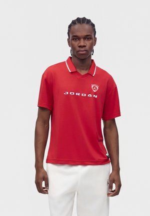 Jeune homme aux cheveux tressés, portant un polo rouge Jordan et un pantalon blanc, debout devant un fond clair uni.
