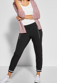 Schwarze, zulaufende Jogginghosen mit seitlichen Reißverschlusstaschen, strukturiertem Bund und gestreiften Bündchen. Kombiniert mit einem weißen Oberteil und einer hellrosa Strickjacke.