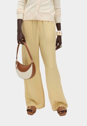 Persoon met beige wijde broek, crèmekleurige vest met knopen, bruine sandalen met gouden gesp, houdt een handtas in twee tinten bruin en crème vast, met grove armbanden.