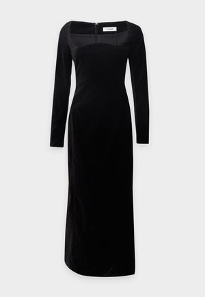 Femme aux longs cheveux foncés portant une robe midi noire ajustée à manches longues et des talons blocs noirs ouverts à l’avant, se tenant devant un fond uni.