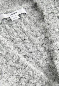 Tela de punto gris con una superficie suave y texturizada, que presenta bucles redondeados. Tag visible con el nombre de la marca "NEXT" en texto negro.