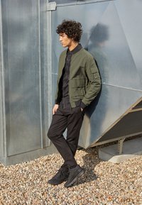 Grüne Bomberjacke, schwarzes Hemd mit Knöpfen, schwarze Hose und schwarze Schuhe. Die Umgebung verfügt über Metallpaneele und einen Kiesboden.