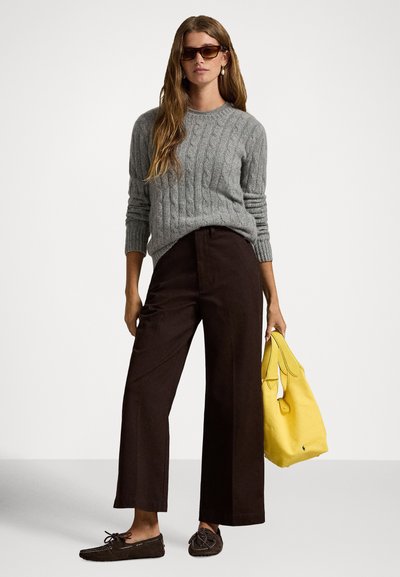 Γυναίκα που φορά καφέ παντελόνι με φαρδιά μπατζάκια, γκρι πουλόβερ πλεκτό με σχέδιο καλώδιο, καφέ μοκασίνια και γυαλιά ηλίου, κρατώντας κίτρινη τσάντα χειρός μπροστά σε απλό φόντο.