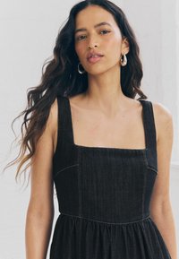 Robe en denim noir avec un décolleté carré, buste ajusté et jupe froncée. Présente des coutures visibles et des bretelles épaisses.