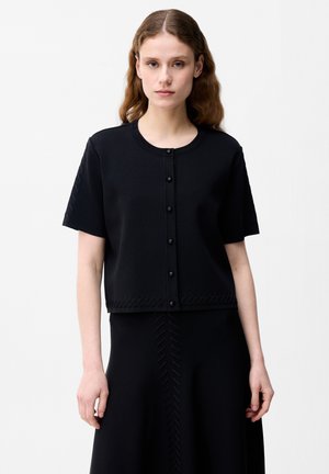 Ung kvinde stående, iført en sort kortærmet knapbluse og matchende sort nederdel med teksturerede chevronmønstre.