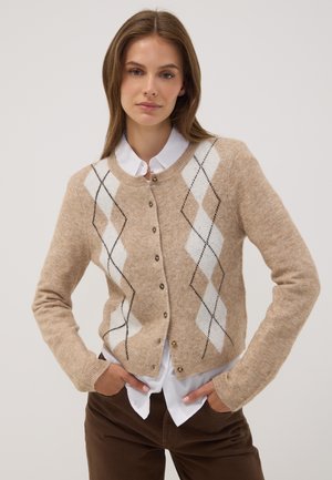 Young woman wearing a beige argyle cardigan over a white shirt and brown pants, standing with hands in pockets against plain background.