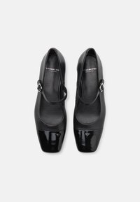 Chaussures en cuir noir avec un embout en vernis noir brillant. Dotées d'une seule sangle réglable avec une boucle en argent et d'une forme carrée arrondie.