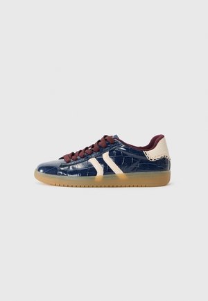 Sneaker basse avec une texture crocodile bleu foncé brillante, patch talon beige, bandes latérales beiges, lacets bordeaux et semelle en gomme.