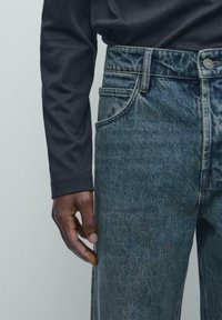 Niebieskie dżinsy z denimu o przecieranej fakturze i pięciokieszeniowym kroju, zestawione z czarną koszulą z długim rękawem. Widoczne przeszycia podkreślają detale.