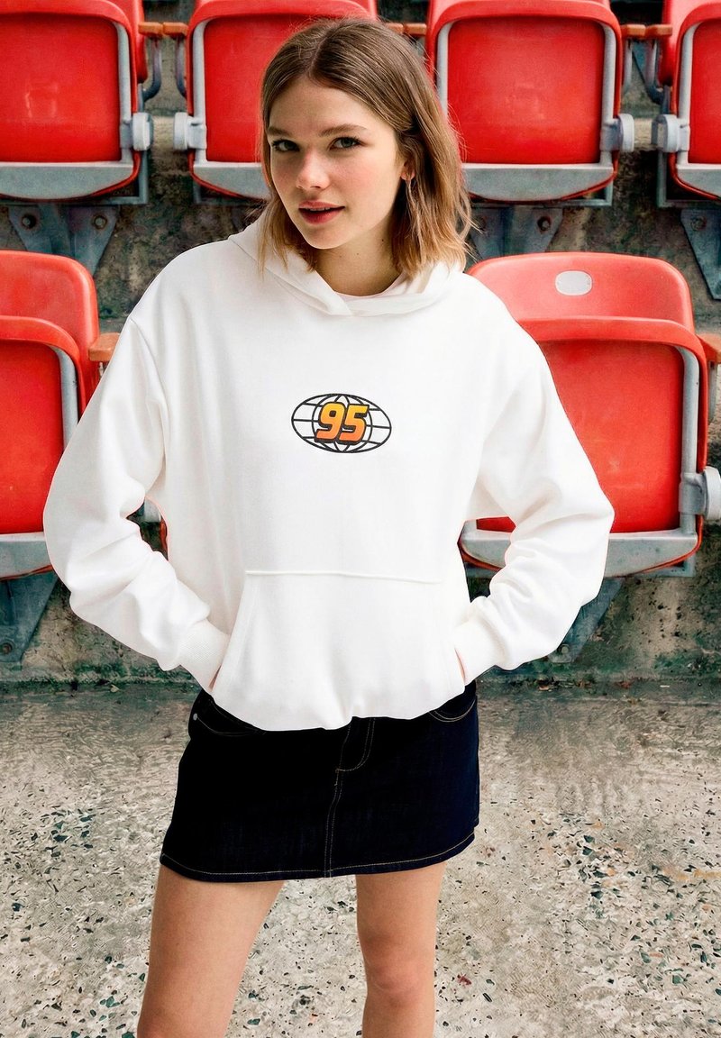 Jeune femme portant un sweat à capuche blanc avec le logo "95" et une jupe noire, debout les mains dans les poches devant des sièges rouges de stade.