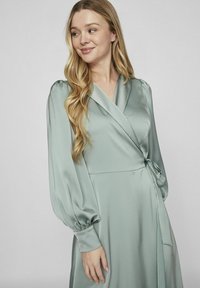 Zelena haljina od satin tkanine u mint nijansi, s dugim napuhanim rukavima i detaljem s vezanjem u struku. Glatka tekstura s suptilnim sjajem.