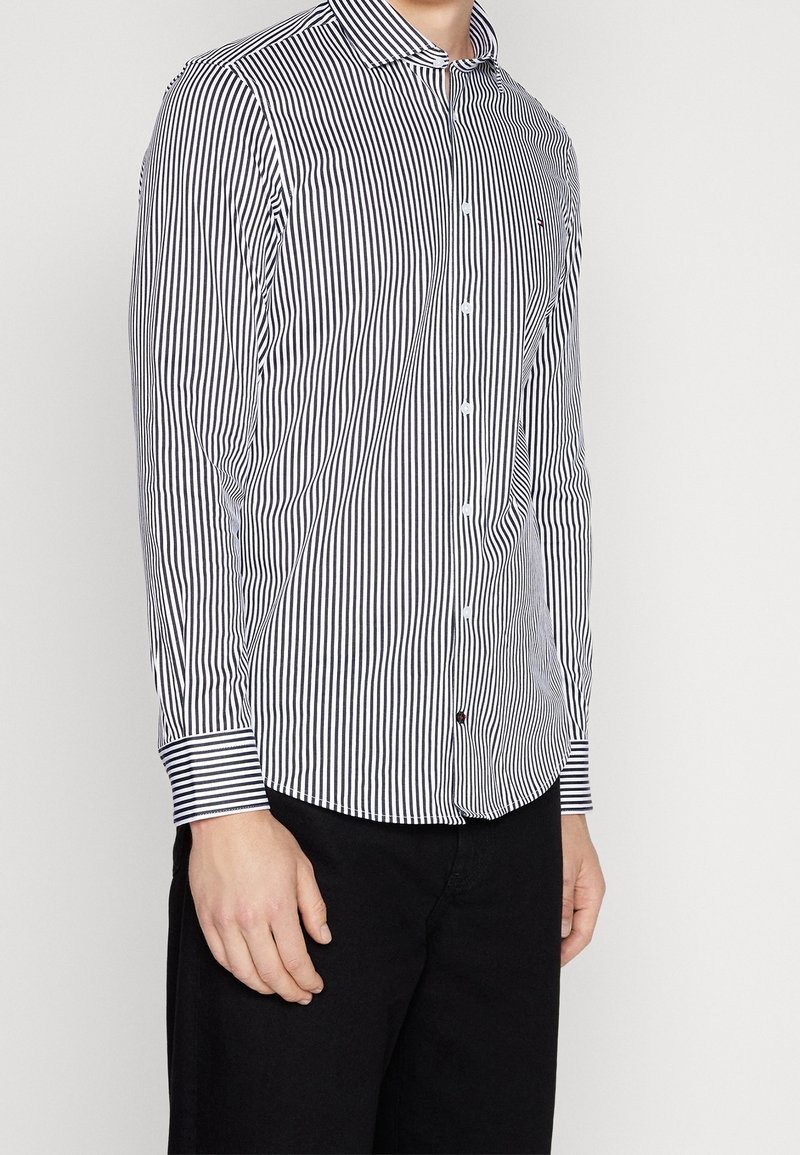 Camicia a maniche lunghe a righe bianche e blu navy con colletto botton-down, orlo curvo e polsini a contrasto. Il materiale sembra liscio con una vestibilità su misura.