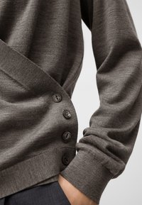 Primo piano di una persona che indossa un cardigan grigio a maglia con quattro bottoni scuri di lato e la mano infilata nella tasca dei pantaloni scuri.