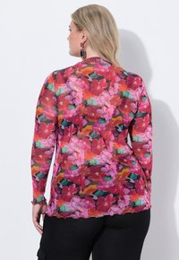 Blusa floreale a maniche lunghe con motivi vibranti di rosso, rosa e verde, realizzata in tessuto trasparente con orlo e polsini scallopati.
