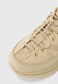 Beige idrettssko med teksturert overdel, elastiske snørebånd, polstret krage og tykk gummisål.