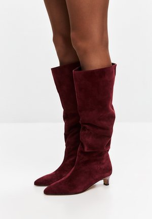 Stivali al ginocchio in suede bordeaux con un design morbido, punta affusolata e un piccolo tacco a blocco. Texture liscia, hardware minimale.