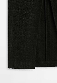 Gros plan d'un tissu tissé noir texturé sur le bord d'un vêtement avec détail de l'ourlet sur fond blanc.
