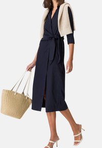 Vestido envolvente azul marino con cintura anudada, presenta un escote en V profundo y mangas de tres cuartos, combinado con un cárdigan de punto beige claro y un bolso de paja.