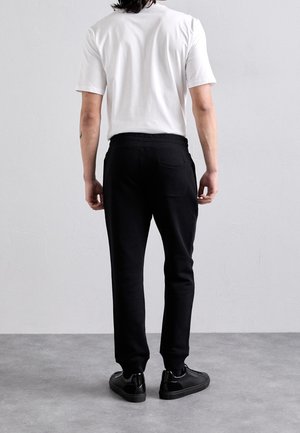 Osoba stojąca tyłem, ubrana w białą koszulkę z krótkim rękawem, czarne spodnie dresowe i czarne trampki na szarej podłodze.