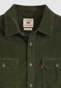 Corduroy shirt in dark green, featuring a pointed collar, button closure, two front pockets, and a small red brand tag on the pocket.