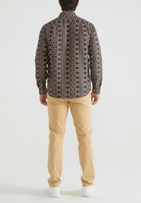 Chemise à manches longues avec un motif géométrique en brun foncé, crème et rouille, associée à un pantalon beige et des baskets blanches.