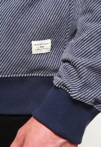 Blue patterned sweatshirt with ribbed navy cuffs and hem. Features a small fabric label with "SELECTED/HOMME" and care instructions.