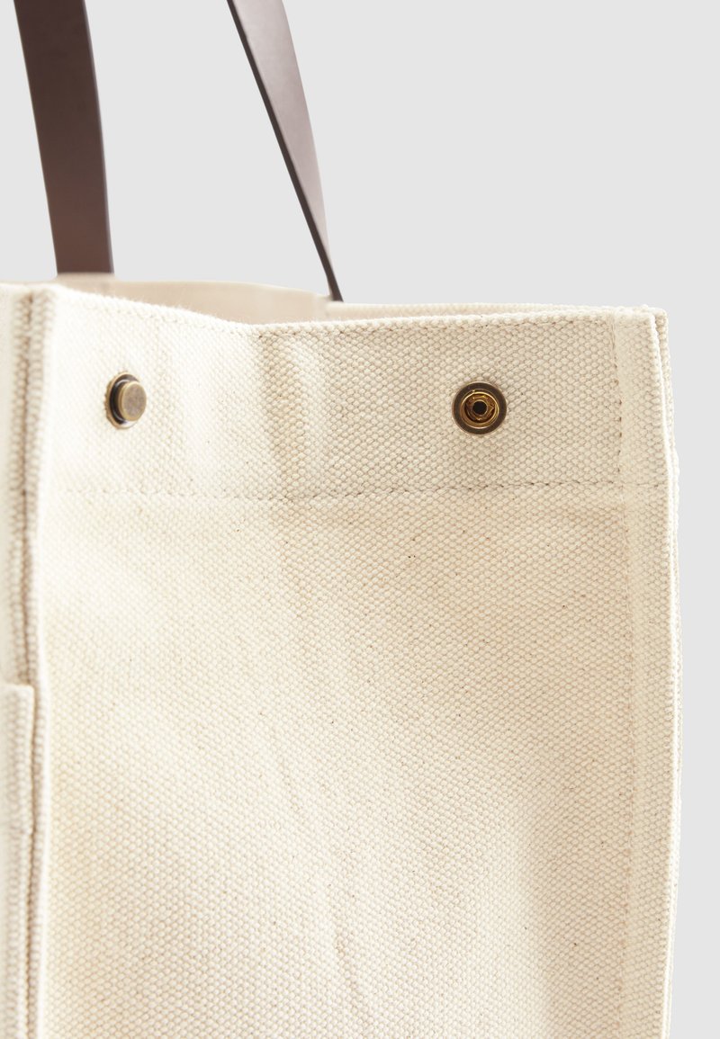 Nahaufnahme einer beige Canvas-Shopper-Ecktasche mit dunkelbraunem Gurt und zwei Metall-Druckknöpfen am oberen Rand vor weißem Hintergrund.