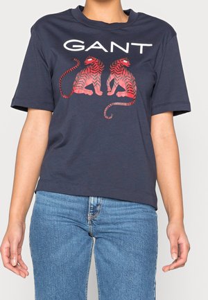 Osoba nosząca granatowy T-shirt z białym napisem "GANT" i dwoma czerwonymi tygrysami zwróconymi twarzami do siebie, w połączeniu z jasnoniebieskimi dżinsami.