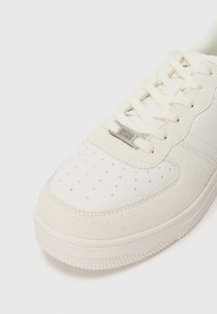 White sneakers with a mix of smooth and suede textures, featuring perforated detailing, flat laces, and a metal logo accent on the side.
