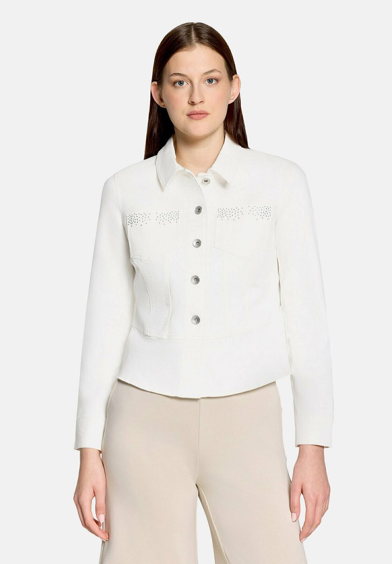 Jeune femme aux cheveux bruns et raides portant une veste blanche boutonnée avec des poches poitrine cloutées et un pantalon beige, debout sur un fond blanc.