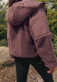 Osoba w fioletowej, strukturalnej polarowej kurtce z pikowanymi i wzmocnionymi ramionami, stojąca na świeżym powietrzu na błotnistej ścieżce w pobliżu skał i drzew.