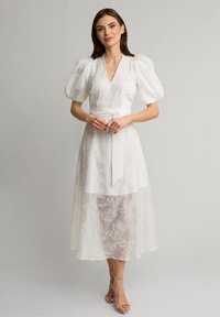 Biała midi sukienka z bufiastymi rękawami, dekoltem w kształcie V i dopasowaną talią. Posiada warstwową, przezroczystą tkaninę z kwiatowymi wzorami oraz szczegół z wiązanym paskiem.