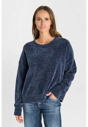 Vrouw met lang bruin haar, die een donkerblauwe gebreide trui en blauwe spijkerbroek draagt, staand met één hand in de zak tegen een effen achtergrond.