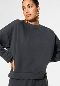 Tummanharmaa ylisuuri huppari, jossa on pyöreä kaula-aukko, ribbaustakin hihnat ja sileä pinta. Yhdistetty suurten koristekorvisten kanssa.