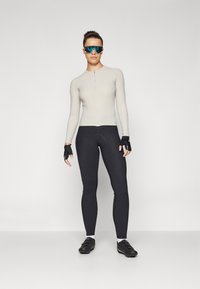 Lichtgrijze lange mouw fietsshirt met een rits aan de voorkant, gecombineerd met zwarte aansluitende leggings en vingerloze zwarte fietshandschoenen.