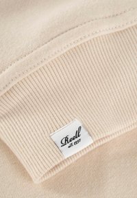 Beige Baumwoll-Sweatshirtkragen mit gerippter Textur und einem rechteckigen Etikett, auf dem der Markenname "Reell" und "est. 1997" in Schwarz gedruckt sind.