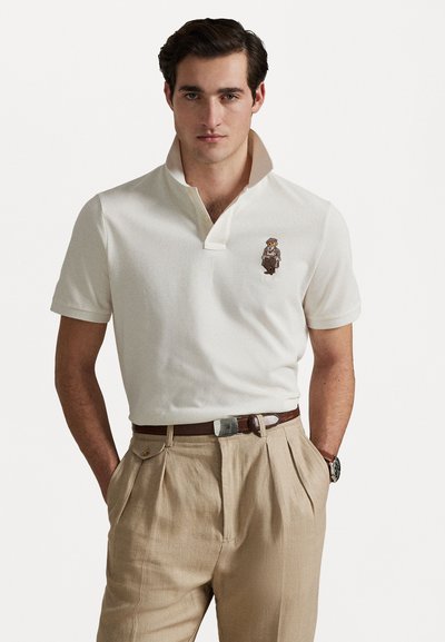 Polo din bumbac alb cu guler împăturit și un personaj brodat pe piept. Asortat cu pantaloni plisati bej deschis și o curea maro.
