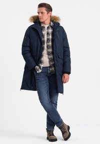 Námořnická modrá péřová bunda s kapucí podloženou kožešinou, s kapsami na zip, nošena přes kostkovanou košili, spárovaná s modrými džíny a hnědými šněrovacími botami.