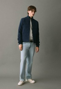 Mladic stoji obučen u tamnoplavu jaknu s patentnim zatvaračem, svijetlosivu majicu, svijetloplave traperice, crni remen i bijele tenisice, a iza njega je jednostavna siva pozadina.