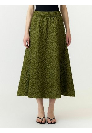 Femme portant une jupe midi verte texturée avec un motif floral et des sandales noires à lanières, debout devant un fond uni.