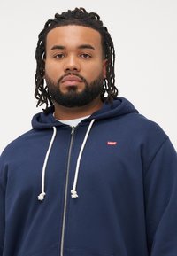 marineblå hettegenser med hvite snorer, med en liten rød Levi's logo. Mykt stoff, avslappet design med en frontlomme.