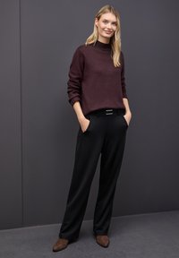 Ung kvinde med blond hår iført en lilla sweater, sorte brede bukser og brune ruskinds støvler, der står med hænderne i lommerne.