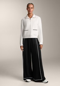 Fehér bomber dzseki fekete cipzárral és zsebkiemelésekkel, laza fekete nadrággal párosítva, amelyek oldalán fehér csíkok találhatók.