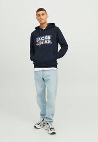 Sudadera navy con un estampado gráfico, bolsillo tipo canguro y capucha con cordón, combinada con jeans de color azul claro y zapatillas en blanco y negro.